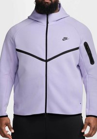 Giacca zip-up Nike viola con accenti neri, caratterizzata da un colletto alto e una tasca con zip sulla manica sinistra, realizzata in tessuto testurizzato.
