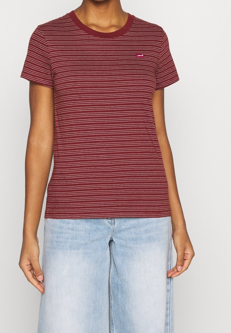 Personne portant un t-shirt à manches courtes rayé bordeaux et blanc et un jean large en denim bleu clair sur un fond uni.