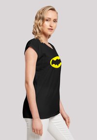 Black t-shirt made of cotton, featuring a large yellow Batman logo on the front. Cap sleeves and a relaxed fit. Paired with white pants.