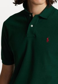 Muž v tmavozeleném polo tričku s červeným vyšívaným logem a dvěma bílými knoflíky u límce.