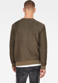 Man med kort hår som bär en olivgrön långärmad tröja och svarta urblekta jeans, stående vänd bort från kameran framför en vit bakgrund.