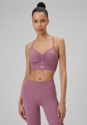 Kobieta z włosami upiętymi w koka, ubrana w pasujący mauve sportowy biustonosz i legginsy z wysokim stanem, stojąca na jednolitym szarym tle.