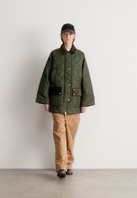 Zöld steppelt dzseki nagy zsebekkel, patentos záródással és magas nyakkal. Világosbarna nadrággal és fekete cipővel párosítva. Egyszerű háttér.