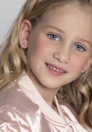 Meisje met golvend blond haar, gekleed in een lichtroze satijnen bomberjack. Ze heeft zilveren studs in haar oren en een gladde huid met subtiele sproeten.