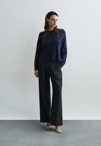 Námornícky pletený sveter s rebrovaným lemom, spárovaný s vysokými, širokými nohavicami v uhlíkovom odtieni a zlatými lodičkami so zakončením na špic. Neutrálny pozadie.