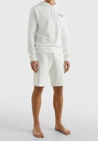 Felpa bianca con cappuccio e pantaloni corti coordinati realizzati in tessuto morbido. Presenta polsini a coste e un piccolo logo sul petto. Nessun motivo o dettaglio.