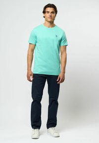 Man står, klädd i en turkos kortärmad t-shirt med liten logo, mörkblå byxor och vita sneakers, mot en enkel ljus bakgrund.