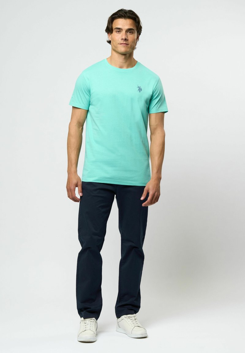 Man står, klädd i en turkos kortärmad t-shirt med liten logo, mörkblå byxor och vita sneakers, mot en enkel ljus bakgrund.