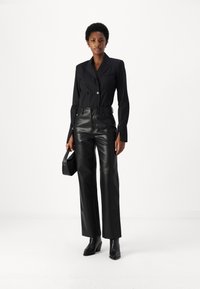 Blazer nero a righe con colletto a contrasto, abbinato a pantaloni in pelle nera a vita alta. La modella tiene una piccola borsa nera e indossa stivali alla caviglia.