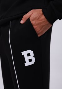 Black sweatpants with white side piping and a large white "B" logo on the left thigh. Soft material and relaxed fit. Hand in pocket detail.