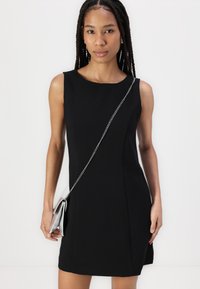 Čierne bezrukávové šaty s okrúhlym výstrihom, priliehavým strihom a hladkou textúrou. V jednej ruke drží striebornú reťazovú crossbody kabelku.