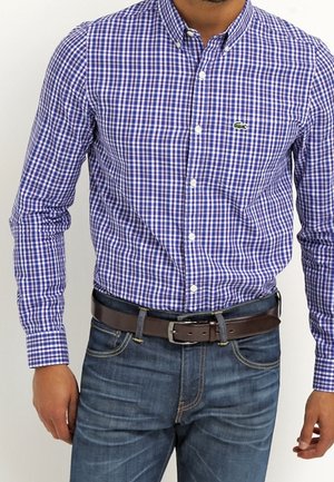 Mężczyzna ubrany w niebiesko-białą koszulę w kratę zapinaną na guziki, wpuszczoną w niebieskie dżinsy z brązowym skórzanym paskiem.