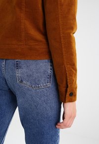 Corduroy jas in gebrand oranje, met een aansluitend model, knoopmanchetten en textuurrijk stof, gecombineerd met blauwe spijkerbroek.