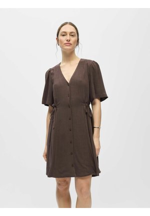 Kvinde iført brun kortærmet skjortekjole med bindebånd i siden, stående mod en ensfarvet hvid baggrund.