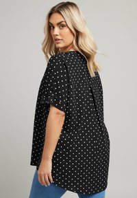 Blusa nera con pois bianchi, maniche corte, vestibilità ampia e dettagli con piega sul retro. Il tessuto sembra liscio. Abbinata a jeans blu.