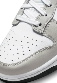 Gros plan d’un bout de basket blanche avec des empiècements en cuir gris, des lacets blancs, des trous de ventilation perforés et des coutures détaillées au-dessus d’une semelle noire.