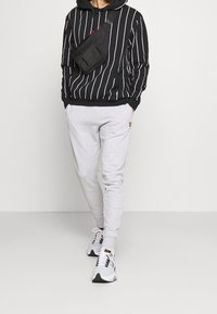 Osoba w czarnym i białym paskowanym hoodie, jasnoszarych dresowych spodniach z małym logo, białych i czarnych trampkach oraz czarnej torbie na ramię.