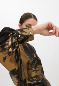 Blusa a maniche lunghe in fantasia tie-dye nero e oro, con maniche a palloncino e bottoni dorati sui polsini. Tessuto liscio e lucente.