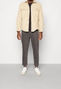 Beżowa koszula na guziki z sztruksu noszona na czarnym t-shircie, w połączeniu z szarymi spodniami i białymi trampkami. Stylizacja jest casualowa i nowoczesna.