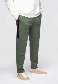 Oliwkowe dresy z czarnymi zamkami w bocznych kieszeniach, charakteryzujące się gładką teksturą i kontrastowymi szwami. W połączeniu z beżowymi sneakersami.