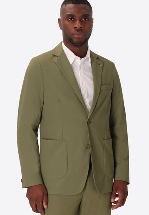 Uomo che indossa una giacca da completo verde oliva e pantaloni abbinati sopra una camicia bianca con bottoni, in piedi davanti a uno sfondo semplice.