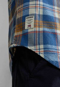 Camisa de cuadros azul, blanca y naranja con una etiqueta de Napapijri Geographic sobre pantalones oscuros de color marino.