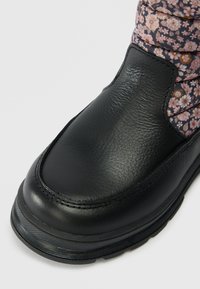 Bota de couro preta com sola grossa e punho de tecido floral em tons de rosa, roxo e castanho, sobre um fundo claro e liso.