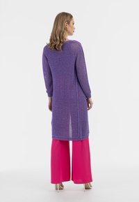 Cardigan lung croșetat violet, cu mâneci lungi, cu fante laterale, asortat cu pantaloni roz cu picioare largi. Material neted și textură tricotată deschisă.