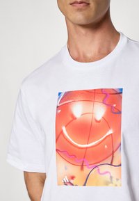 Man in een wit T-shirt met een kleurrafische basketbalafbeelding die een glimlachend gezicht en abstracte paarse en roze lijnen toont.