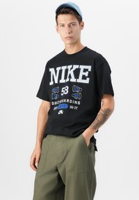 Juodas medvilnės marškinėliai su dideliu baltu "NIKE" užrašu ir spalvingu grafiniu piešiniu. Derinami su žaliais, laisvo kirpimo cargo džinsais.