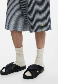 Fekete keresztpántos szandál, matt felülettel, krém színű bordázott zoknival párosítva, csíkos sötétkék sort alatt, melyen kis sárga logó részlet található.