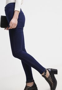 Tmavě modré skinny džíny vyrobené z denimu s středním pasem, mají hladkou texturu a jemné švy. Na nohou jsou černé sandály na podpatku.