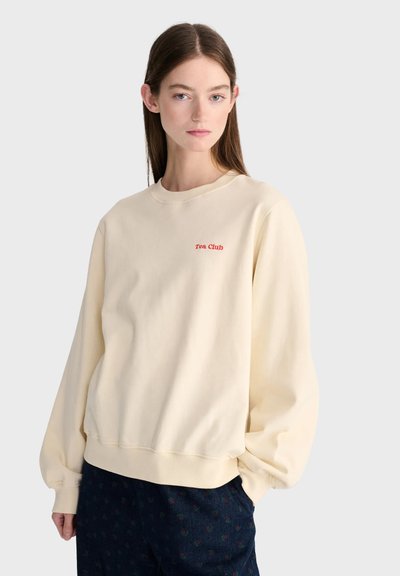 Jeune femme aux cheveux bruns raides portant un sweat crème avec le texte "Tea Club" et un pantalon foncé à motifs sur un fond uni.