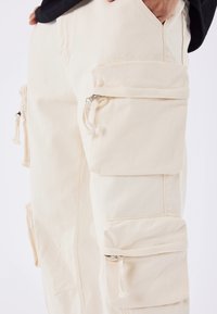 Pantalones cargo de algodón beige claro, con múltiples bolsillos con cremallera, un ajuste relajado y detalles de costura visibles.