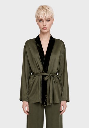 Femme aux cheveux blonds courts portant une robe en satin vert olive avec bordure noire nouée à la taille, debout devant un fond clair uni.