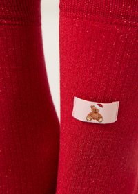 Chaussettes rouges en maille avec une finition texturée et des paillettes discrètes. Elles comportent une étiquette rose représentant un ours en peluche portant un chapeau.