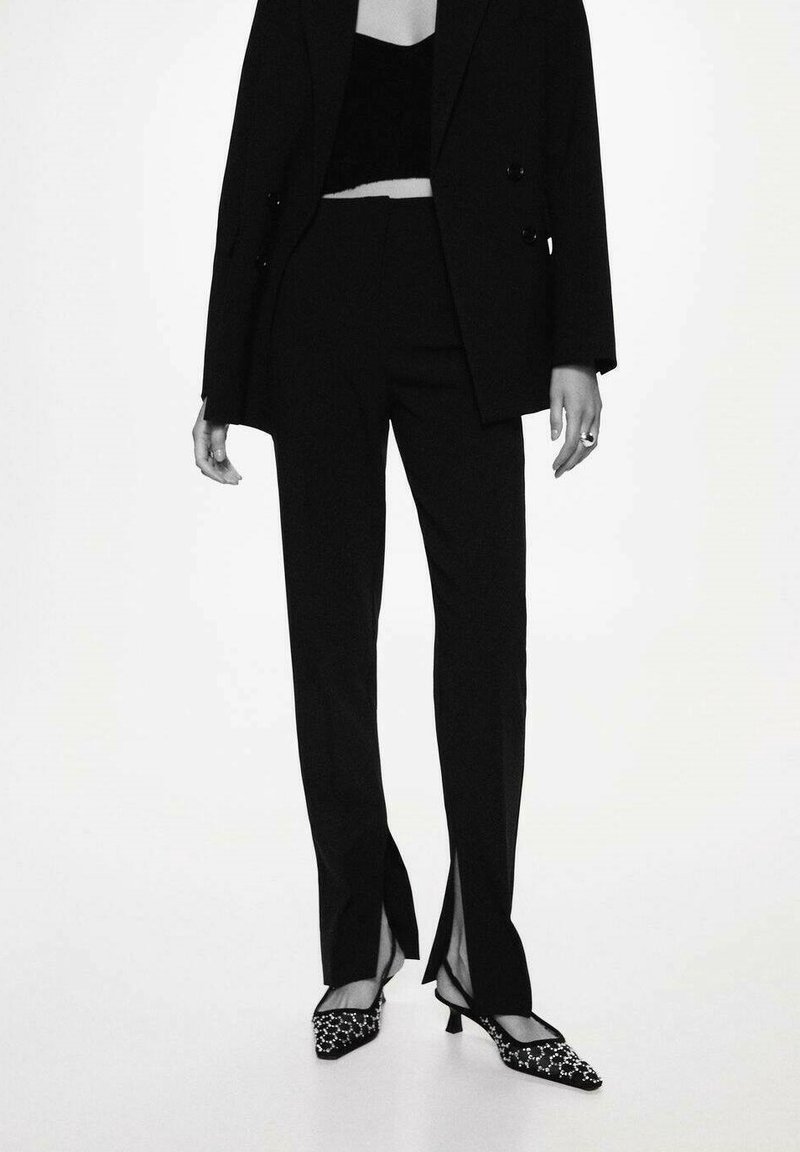 Veste de costume noire sur mesure et pantalon taille haute avec fentes latérales. Talons texturés à motifs noirs et blancs complètent la tenue.