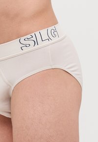 Beige Baumwollslips mit einem weißen Bund und navyfarbener Schrift. Der Stoff hat eine weiche Textur und ein einfaches Design.