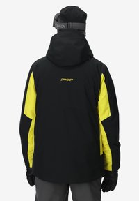 Schwarze und gelbe Skijacke mit Kapuze, aus wasserdichtem Material. Hat ein Logo auf dem Rücken und schwarze Handschuhe, die von der Person getragen werden.