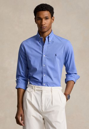 Άνδρας με ανοιχτό μπλε πουκάμισο με κουμπιά και λογότυπο, με τα μανίκια σηκωμένα, λευκός πλισέ παντελόνι και ρολόι χειρός, μπροστά σε απλό μπεζ φόντο.