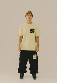 Giovane uomo con tatuaggi sta in piedi di fronte indossando una maglietta con logo giallo pallido e pantaloni neri larghi con logo abbinato, scarpe da ginnastica nere.