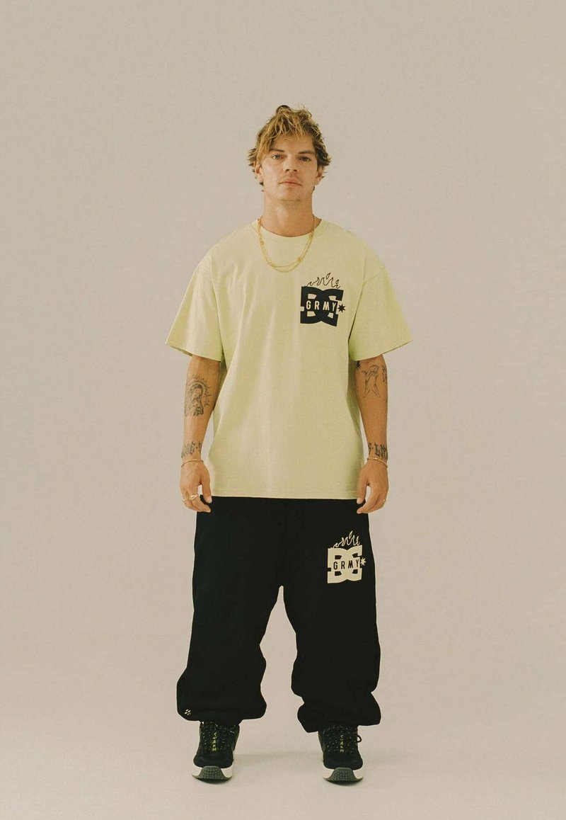 Giovane uomo con tatuaggi sta in piedi di fronte indossando una maglietta con logo giallo pallido e pantaloni neri larghi con logo abbinato, scarpe da ginnastica nere.