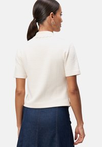 Kortærmet hvid top med et struktureret mønster og en bølget krave, stylet med en denimnederdel. Har en tætsiddende silhuet.