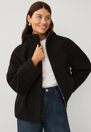 Kvinde smiler mens hun har en sort tekstureret lynlåstrøje, hvid top, blå jeans og gyldne hoop-øreringe på foran en ensfarvet baggrund.