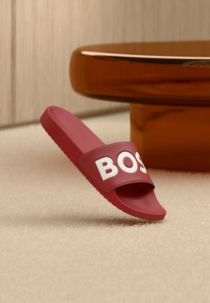 Rote Rutsche-Sandale mit dickem Riemen, der teilweise weiße Buchstaben "BOS" zeigt, auf beigem Teppich neben einem runden, bernsteinfarbenen Tisch platziert.