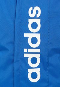 Niebieska tkanina o gładkiej fakturze z dużym, białym logo "adidas" wyraźnie wyeksponowanym. Projekt zawiera widoczne detale ściegów.