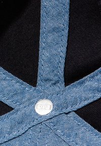 Les salopettes en jean présentent des bretelles croisées dans le dos avec un bouton-pression argenté, un tissu bleu texturé et un tissu noir contrastant en dessous.