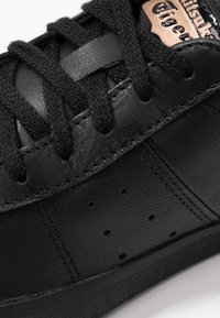 Gros plan sur une basket en cuir noir avec des lacets noirs, un panneau latéral perforé et une étiquette de marque partiellement visible en beige et noir sur la languette.