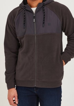 Hombre con sudadera de forro polar gris oscuro con cremallera y cordones negros, y vaqueros azul oscuro, de pie con una mano en el bolsillo.