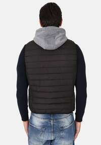 Gilet imbottito nero con design a righe orizzontali, indossato sopra un felpa con cappuccio grigia. Jeans in denim blu visibili nella parte inferiore.